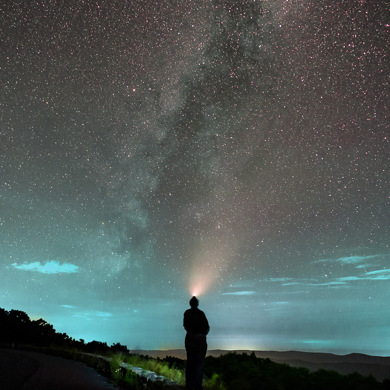 Person with headlamp stands by a road under a starry sky and the Milky Way.